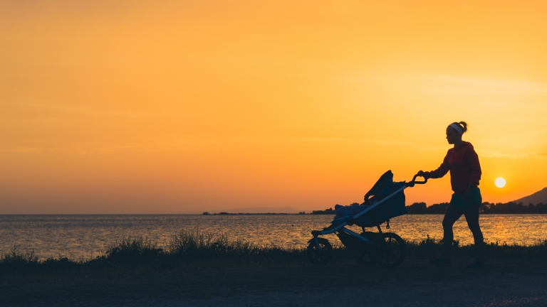 Mother walking on beach with stroller enjoying motherhood at sunset landscape. Jogging or power walking woman with pram. Beautiful inspirational coastline landscape.
